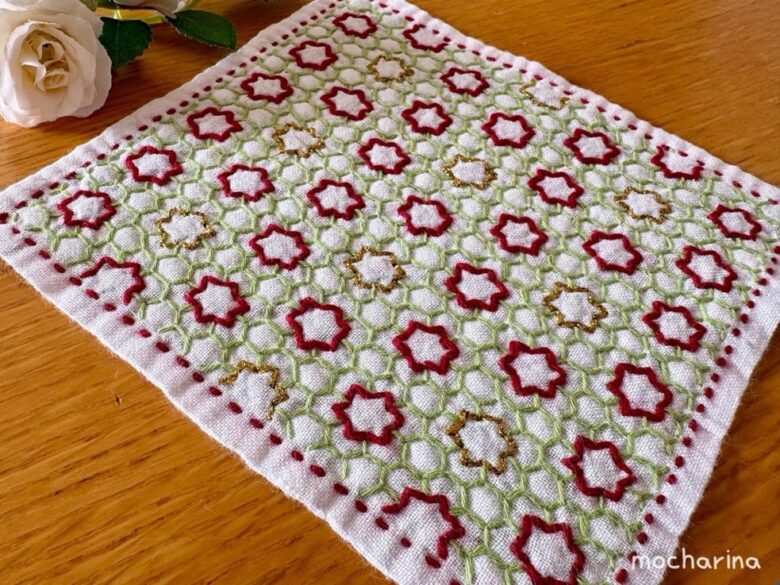 刺し子ふきん「変わり亀甲」を斜め撮りした完成写真。緑の亀甲柄に赤と金色の星型がきらめいて見えるデザイン。